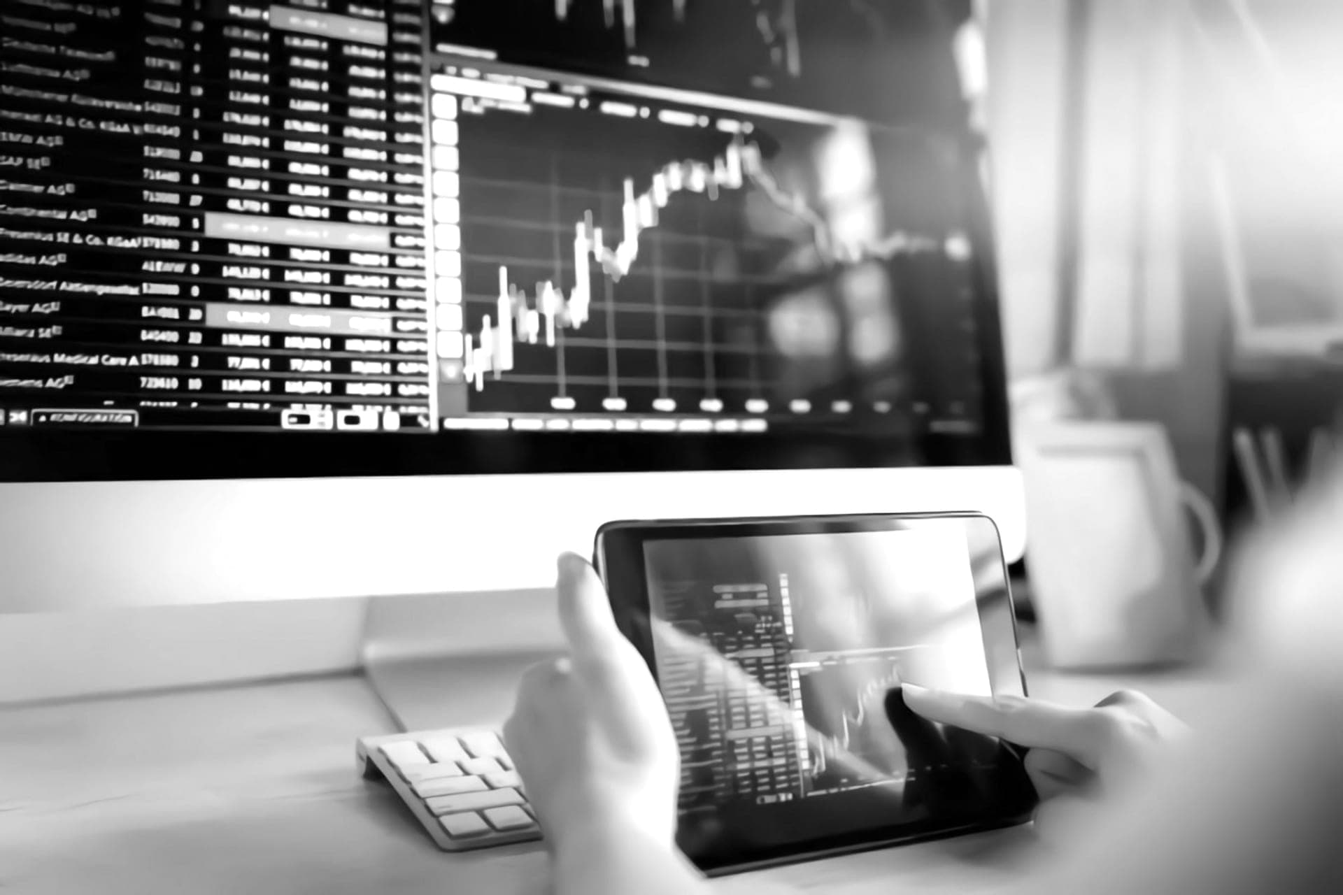 Stock-market charts on a monitor and on a tablet held in one hand, in black and white