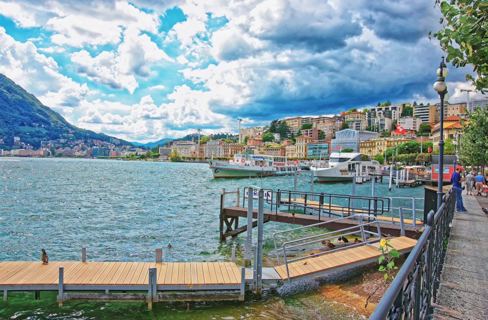 A lakeside town below wooded mountains, with boats moored along the shore
